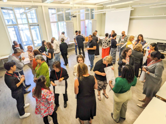 A large group of people stands in small clusters, talking and engaging in discussion inside a bright, modern room with large windows and a projector screen. The setting suggests a workshop or networking event, with participants holding papers and actively interacting.